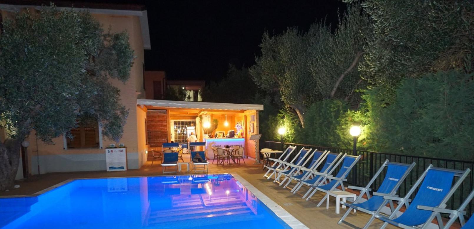 Piscina illuminata di notte con sedie a sdraio e bar sullo sfondo.