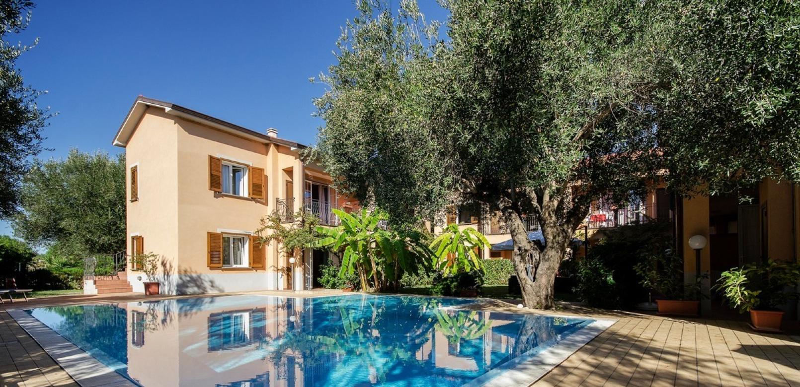 Villa con piscina, giardino rigoglioso, alberi e cielo azzurro.