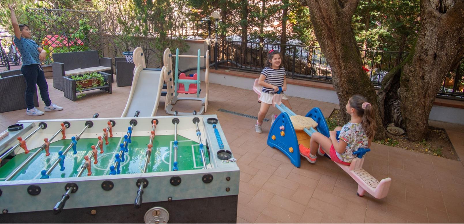 Bambini giocano all'aperto con altalena, scivolo e calcio balilla in giardino.