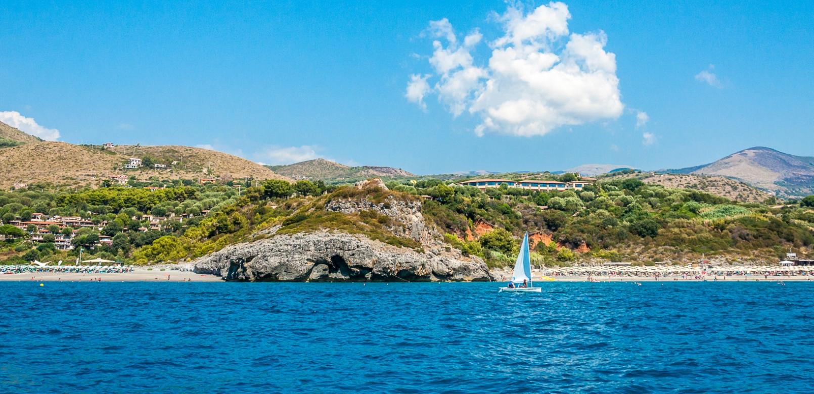 Spiaggia con barca a vela, colline verdi e cielo azzurro.