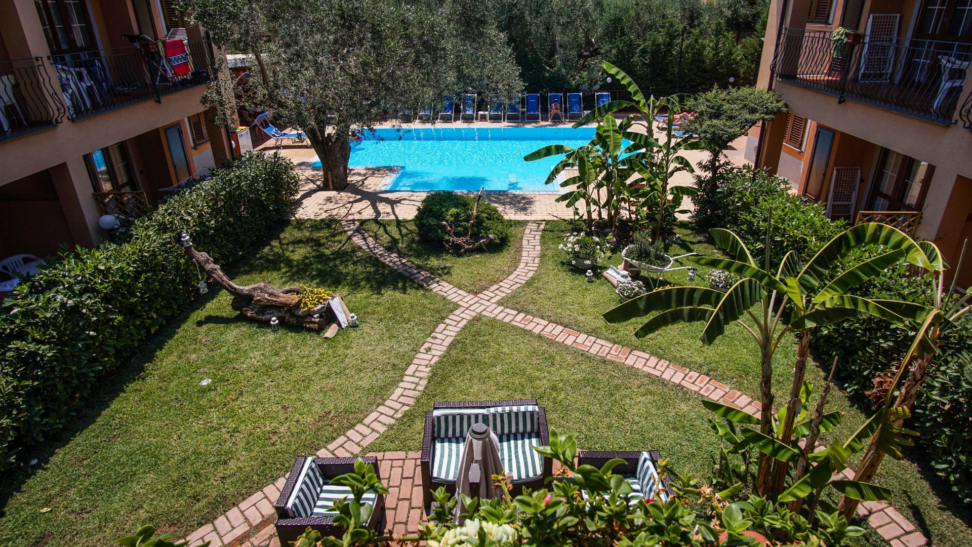 Giardino con piscina, sentieri in mattoni e vegetazione lussureggiante, circondato da edifici.