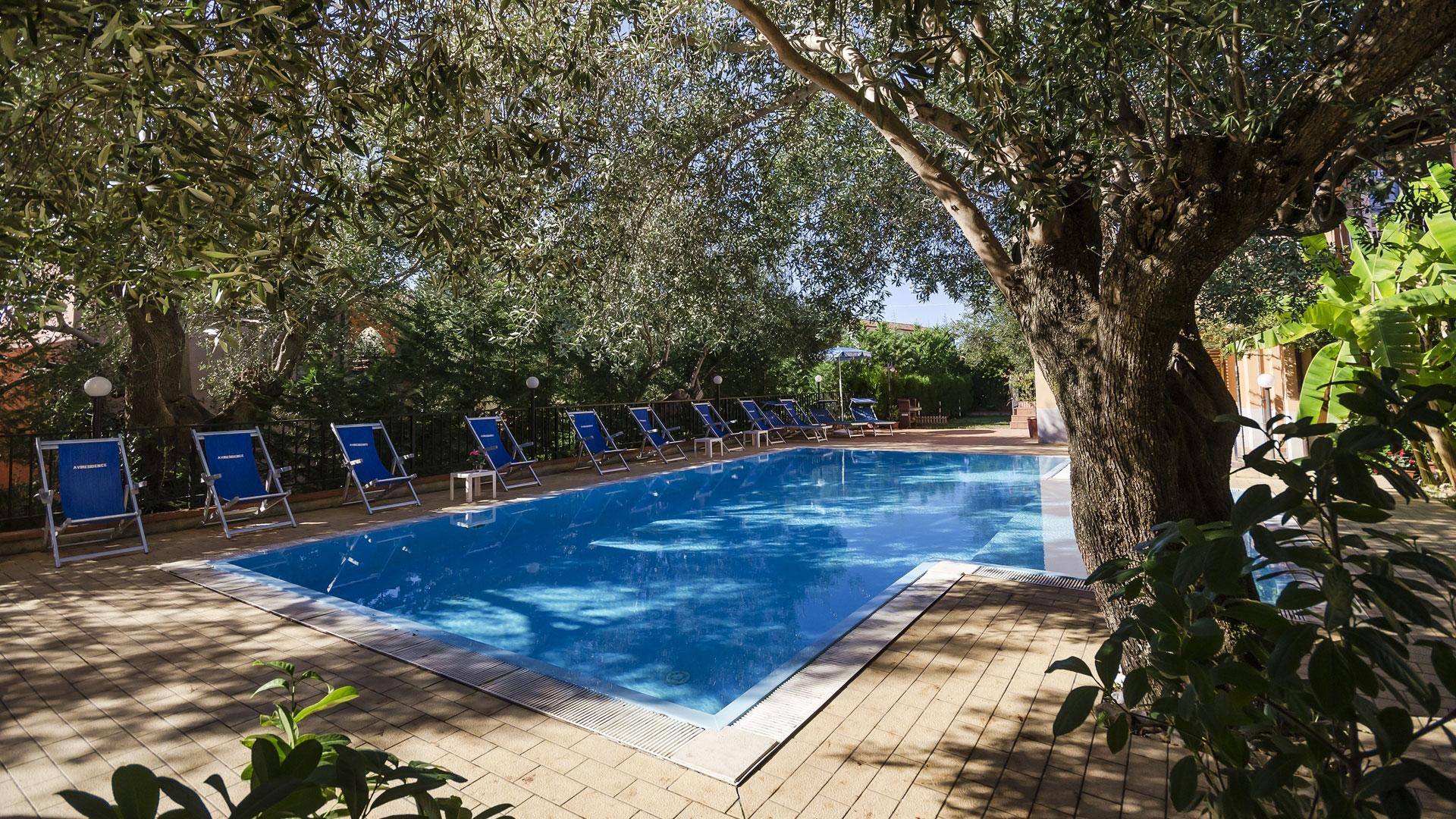 Piscina all'aperto con lettini blu, circondata da alberi e vegetazione rigogliosa.