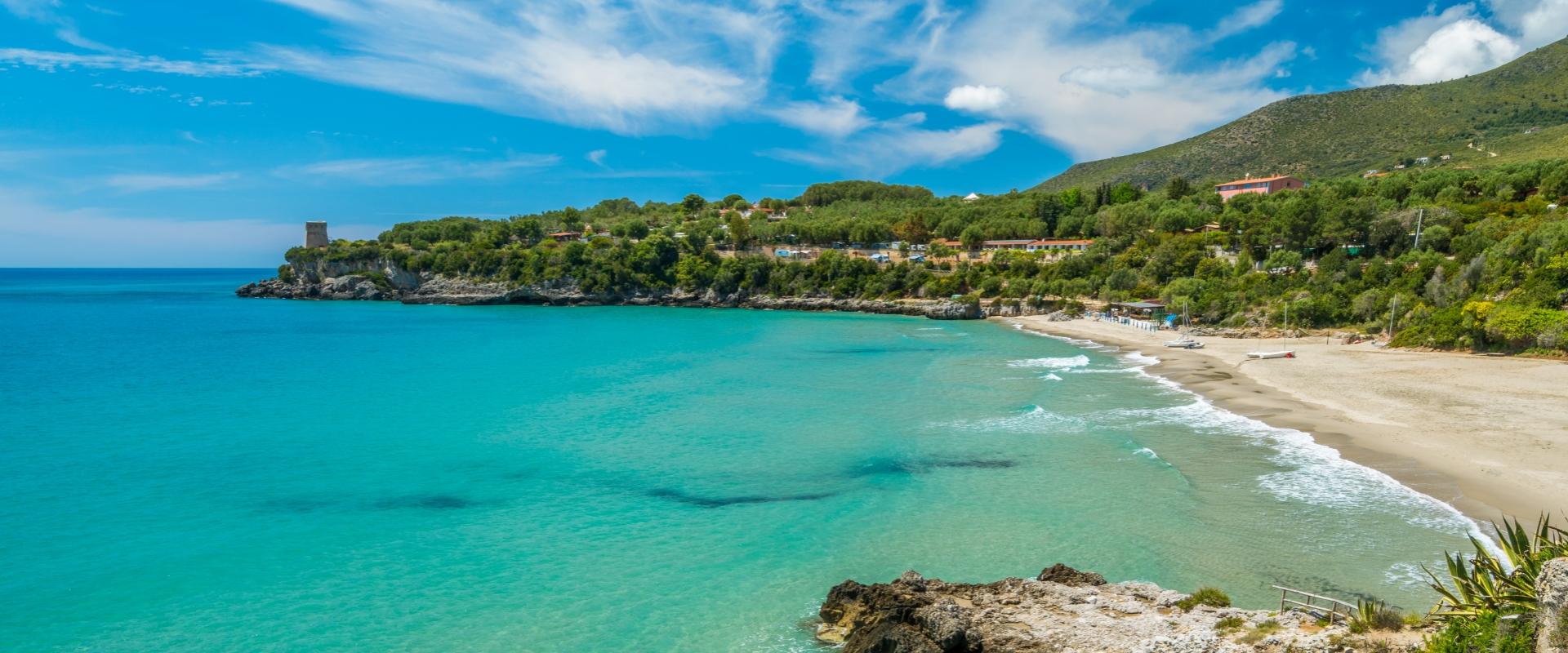 Spiaggia con mare turchese, scogliera e vegetazione lussureggiante sotto un cielo azzurro.