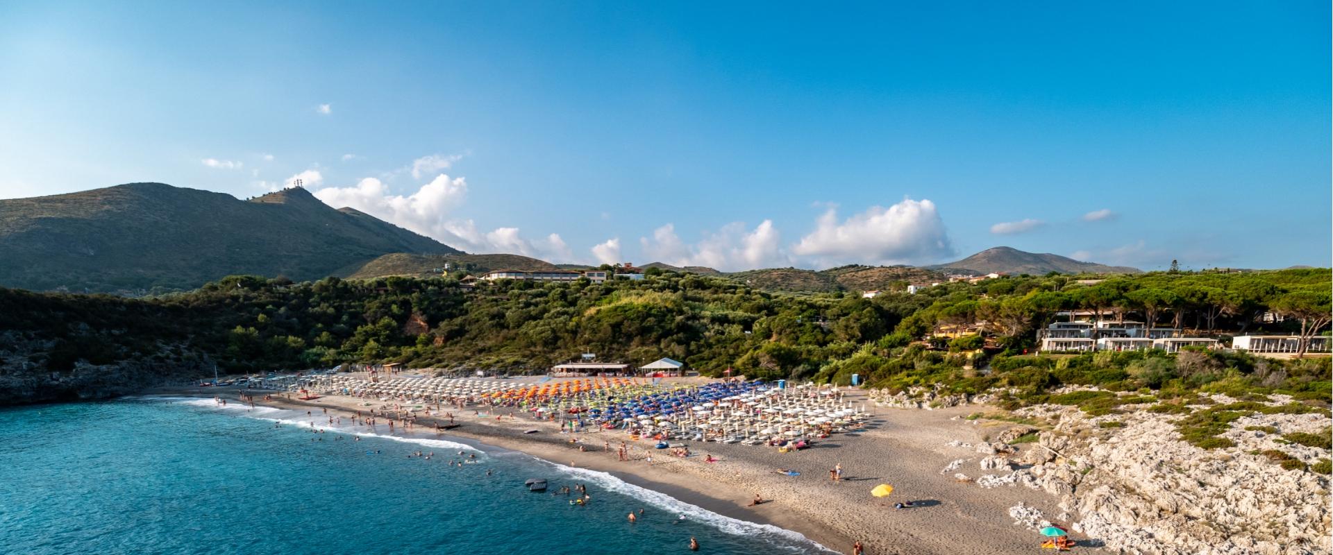 Spiaggia affollata con ombrelloni colorati, mare azzurro e colline verdi sullo sfondo.