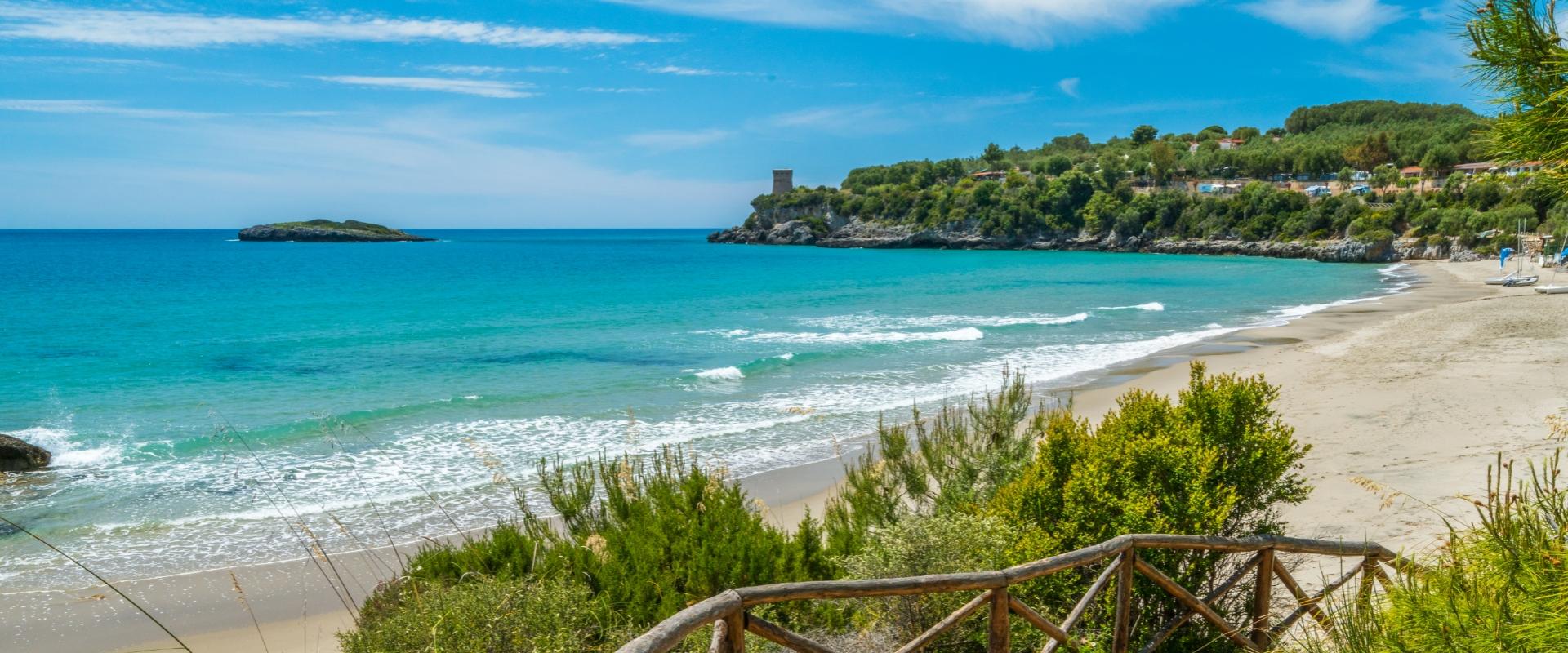 Spiaggia sabbiosa con mare turchese e torre su promontorio verde.