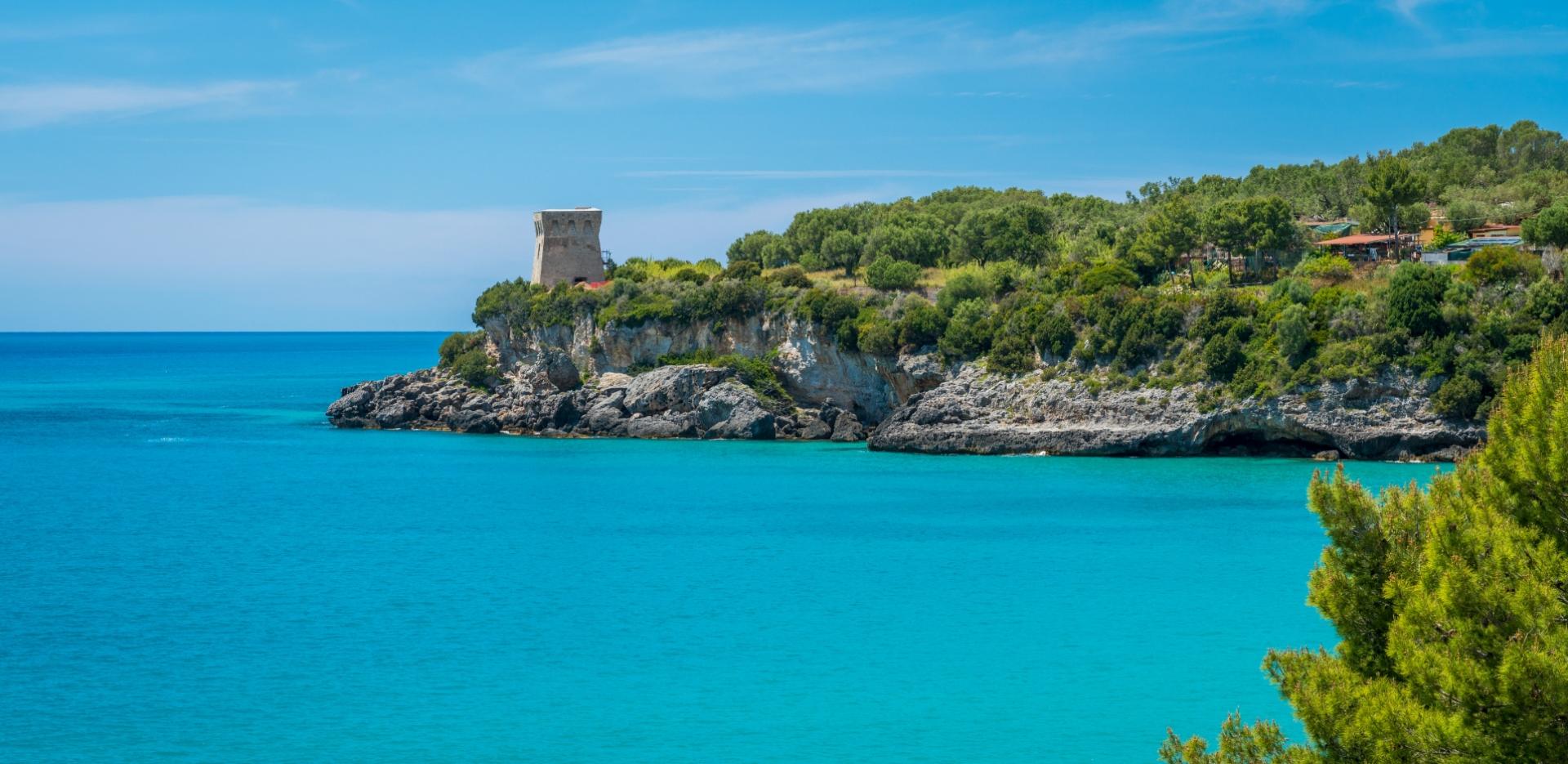 Torre costiera su scogliera verdeggiante, affacciata su mare turchese e cielo azzurro.