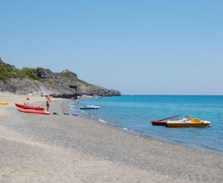 Spiaggia con mare cristallino, pedalò colorati e pochi bagnanti, circondata da scogliere.