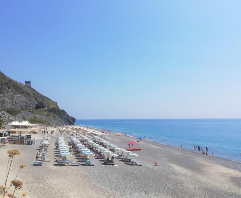 Spiaggia con ombrelloni colorati, mare azzurro e colline verdi sullo sfondo.