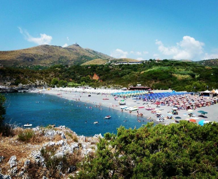 Spiaggia affollata con ombrelloni colorati, mare blu e colline sullo sfondo.