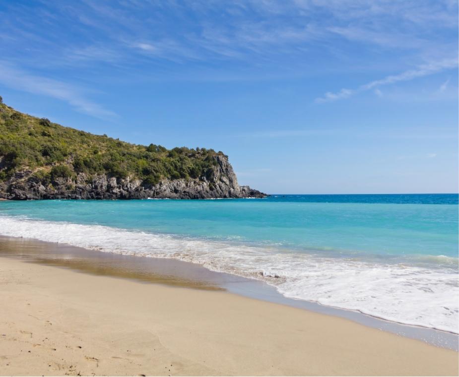 Spiaggia sabbiosa con mare turchese e promontorio verde sotto un cielo limpido.