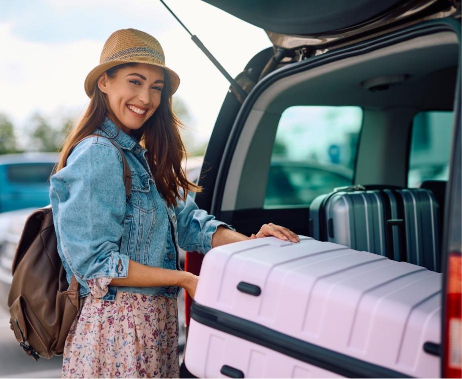 Donna sorridente carica una valigia nel bagagliaio di un'auto per un viaggio.