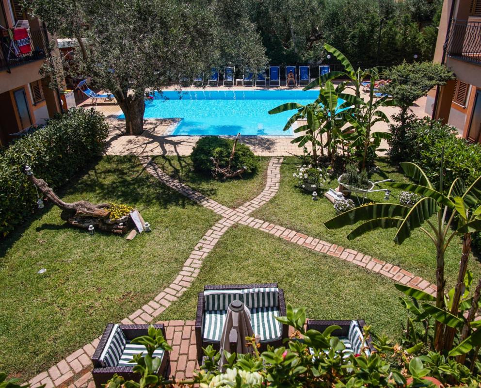 Giardino con piscina, sentieri in mattoni e vegetazione lussureggiante, circondato da edifici.